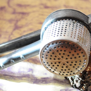 May include: A silver potato masher with a black handle. The masher has a round, perforated head with a star-shaped pattern.
