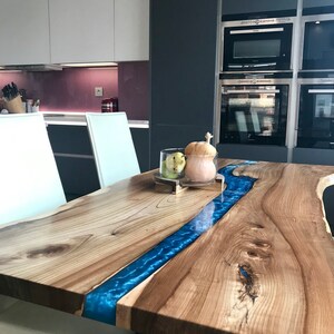 May include: A wooden dining table with a blue epoxy resin river running down the center. The table has a natural wood finish and is supported by a black metal base. Two white chairs are visible in the foreground.