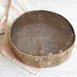 May include: A round, wooden flour sifter with a wire mesh screen. The sifter is brown and has a worn, rustic appearance.