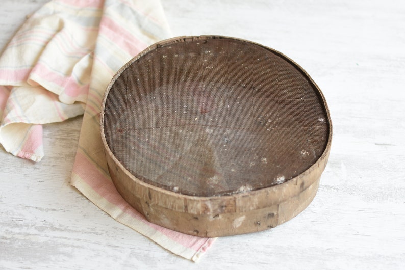 May include: A round, wooden flour sifter with a wire mesh screen. The sifter is brown and has a worn, rustic appearance.