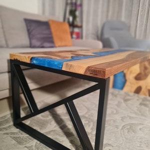 May include: A wooden coffee table with a blue epoxy river design and a black metal geometric base. The table has the text "RIVULETT" on the surface.