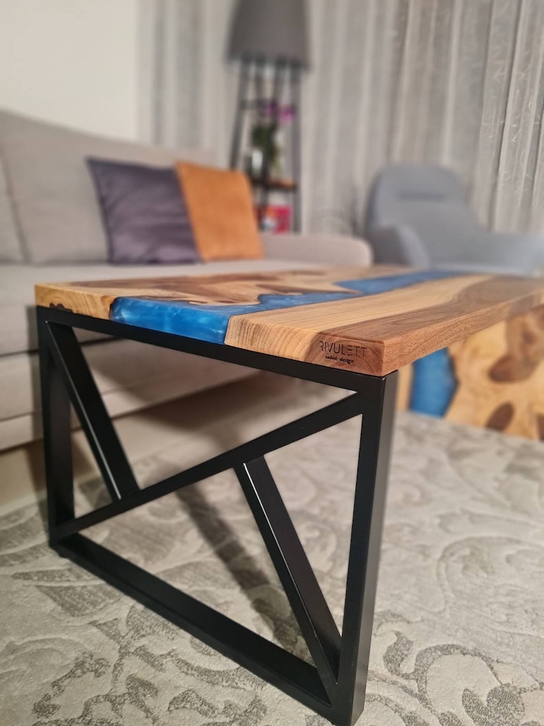May include: A wooden coffee table with a blue epoxy river design and a black metal geometric base. The table has the text "RIVULETT" on the surface.