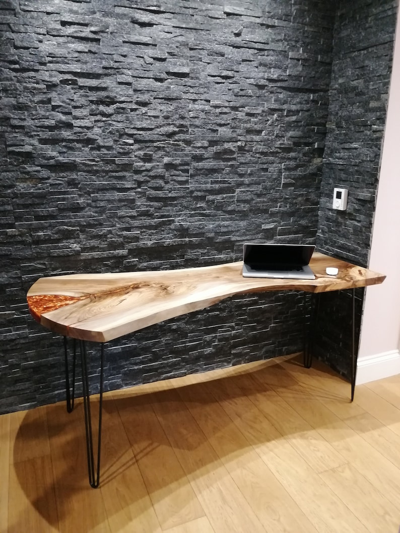 Pu&ograve; includere: Una scrivania in legno a bordo vivo con una finitura in legno naturale e un riempimento in resina epossidica trasparente. La scrivania ha una forma unica e organica ed &egrave; sostenuta da quattro gambe a forcina nere. La scrivania &egrave; posizionata contro un muro di piastrelle di ardesia nera.