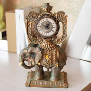 Può includere: Un orologio dorato con un design elaborato si trova sopra una statuetta di elefante. Il quadrante dell'orologio ha numeri romani e una cornice dorata.