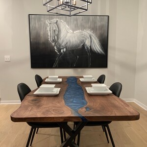 May include: A large wooden dining table with a blue resin river running down the center. The table has a live edge and is supported by a black metal X-shaped base. There are six black chairs around the table, and a painting of a white horse hangs on the wall behind the table.