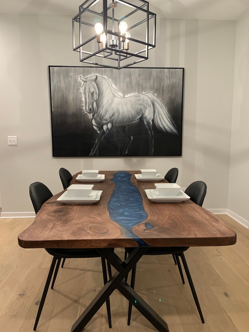 May include: A large wooden dining table with a blue resin river running down the center. The table has a live edge and is supported by a black metal X-shaped base. There are six black chairs around the table, and a painting of a white horse hangs on the wall behind the table.