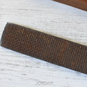 May include: Close-up of a well-used, rectangular rasp file with a textured, diamond-patterned surface. The file is a rusty brown color and appears to be made of metal. The background is a white wooden surface.