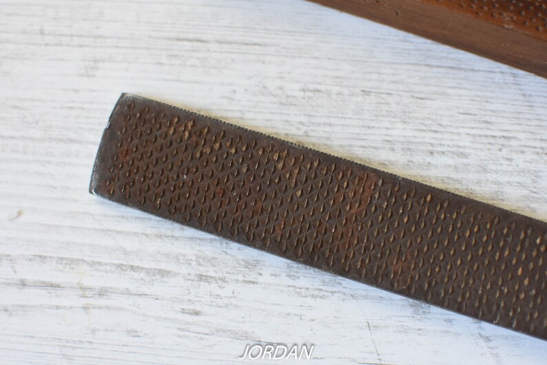 May include: Close-up of a well-used, rectangular rasp file with a textured, diamond-patterned surface. The file is a rusty brown color and appears to be made of metal. The background is a white wooden surface.