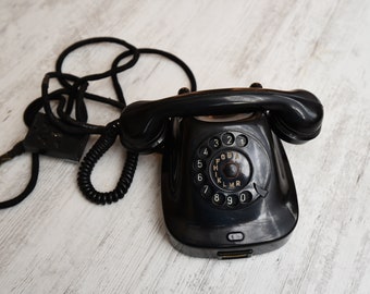 May include: A black rotary dial phone with a coiled cord and a black handset. The phone has a circular dial with numbers 1 through 0 and letters A through R.