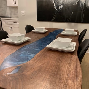 May include: A large wooden dining table with a blue epoxy river running down the center. The table has a live edge and is made of walnut wood. There are four white bowls and plates on the table.