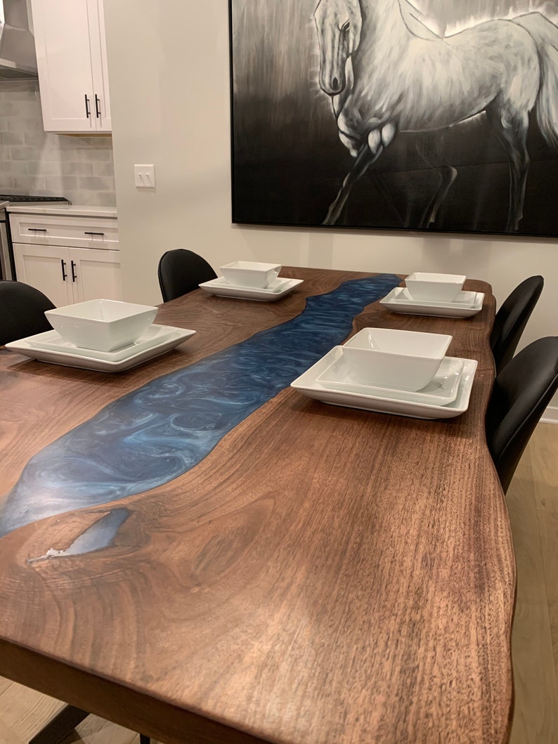 May include: A large wooden dining table with a blue epoxy river running down the center. The table has a live edge and is made of walnut wood. There are four white bowls and plates on the table.