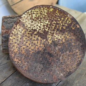Puede incluir: Una cabeza de regadera redonda de metal oxidada con un acabado de pintura dorado y un patr&oacute;n de peque&ntilde;os agujeros. La cabeza de la regadera est&aacute; sobre una superficie de madera.