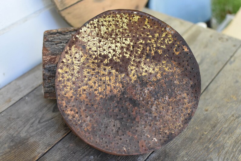 Puede incluir: Una cabeza de regadera redonda de metal oxidada con un acabado de pintura dorado y un patr&oacute;n de peque&ntilde;os agujeros. La cabeza de la regadera est&aacute; sobre una superficie de madera.