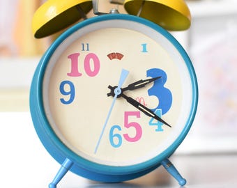 Puede incluir: Un despertador azul con campanas amarillas y una esfera blanca. El reloj tiene agujas negras y números de colores. Son las 10:55.