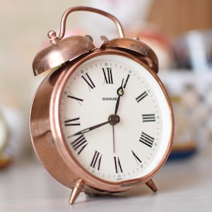 Op de afbeelding: Een koperkleurige wekker in vintage-stijl met een witte wijzerplaat en zwarte Romeinse cijfers. De klok heeft een klassiek belontwerp en staat op drie kleine voetjes. De wijzerplaat leest "BAYARD".