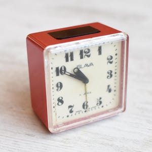 Puede incluir: Un reloj despertador rojo con una esfera blanca y números negros. El reloj dice "Slava" y tiene una aguja negra que apunta al número 12.