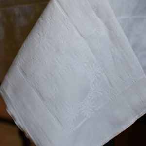 May include: White linen tablecloth with an embossed floral pattern. The tablecloth is folded in half and draped over a table.