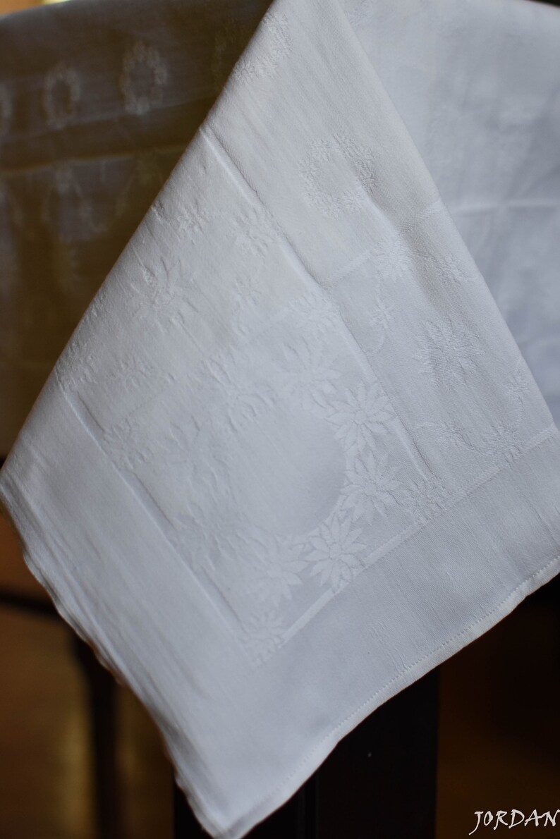 May include: White linen tablecloth with an embossed floral pattern. The tablecloth is folded in half and draped over a table.