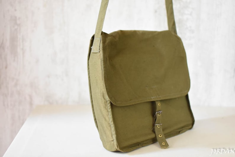 May include: A green canvas messenger bag with a flap closure and a buckle strap. The bag has a worn look and is made of a durable material.