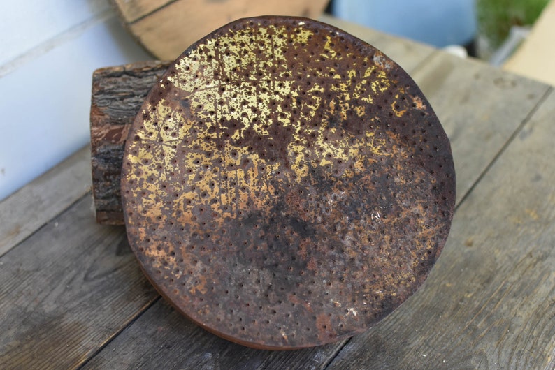 Puede incluir: Una cabeza de regadera redonda de metal oxidada con un acabado de pintura dorado y un patr&oacute;n de peque&ntilde;os agujeros. La cabeza de la regadera est&aacute; sobre una superficie de madera.