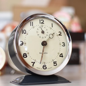 May include: A round analog alarm clock with a polished chrome frame and a cream-colored face. The dial features black Arabic numerals, a small sub-dial at the top, and thin hands. The text "BAYARD" and "MADE IN FRANCE" is printed on the face.