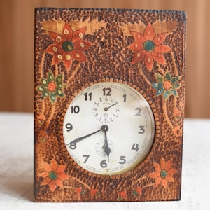May include: A vintage rectangular clock with a brown, textured exterior and floral designs in orange, green, and red. The clock face is white with black numerals and hands. A small dial is located above the center.