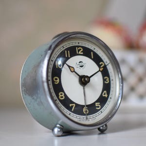 May include: A vintage, round alarm clock with a light blue metal casing. The clock face is white with black and gold numerals, and black hands. The clock has a small, round base and a silver-colored bezel.