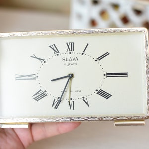 May include: A gold-coloured rectangular alarm clock with Roman numeral markings on the face. The clock face is white with black markings and the text "SLAVA 11 jewels" and "MADE IN USSR".