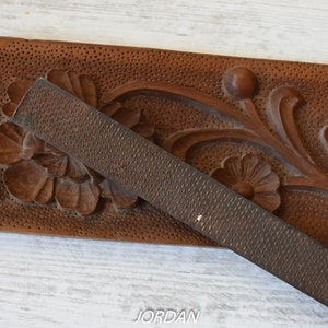 May include: A close-up of a wooden carving with floral designs and a metal rasp. The wood is a warm brown color, and the rasp is a dark gray. The carving features detailed flowers and leaves.