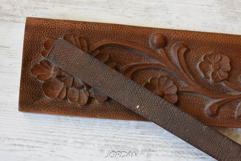 May include: A close-up of a wooden carving with floral designs and a metal rasp. The wood is a warm brown color, and the rasp is a dark gray. The carving features detailed flowers and leaves.