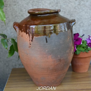 Può includere: Un grande vaso di ceramica marrone smaltato con due manici. Il vaso ha un aspetto rustico e invecchiato ed è appoggiato su un tavolo di legno.