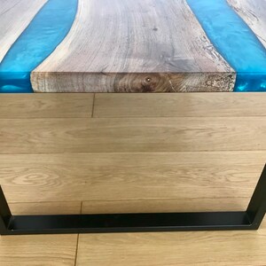 Puede incluir: Una mesa de caf&eacute; de madera con un marco de metal negro. La mesa tiene un acabado de madera natural con un r&iacute;o de resina epoxi azul que la atraviesa.