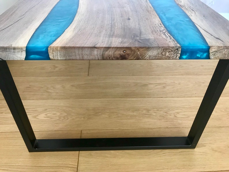 Puede incluir: Una mesa de caf&eacute; de madera con un marco de metal negro. La mesa tiene un acabado de madera natural con un r&iacute;o de resina epoxi azul que la atraviesa.
