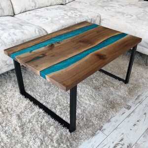 Puede incluir: Una mesa de centro de madera con una incrustaci&oacute;n de resina epoxi azul turquesa. La mesa tiene un marco de metal negro y est&aacute; sentada sobre una alfombra de pelo blanco.