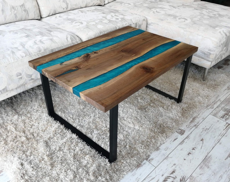 Puede incluir: Una mesa de centro de madera con una incrustaci&oacute;n de resina epoxi azul turquesa. La mesa tiene un marco de metal negro y est&aacute; sentada sobre una alfombra de pelo blanco.