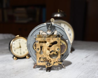 May include: A vintage alarm clock with the back removed, revealing the inner workings of the clock's mechanism. The clock has a silver-colored case and gold-colored gears.