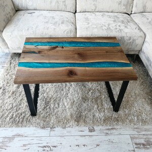 Puede incluir: Una mesa de centro de madera con una incrustaci&oacute;n de resina epoxi turquesa. La mesa tiene un marco de metal negro y est&aacute; sentada sobre una alfombra blanca.