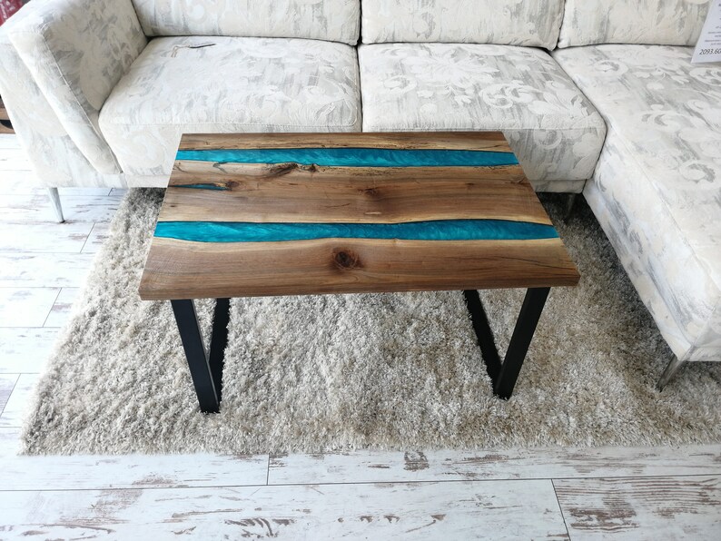 Puede incluir: Una mesa de centro de madera con una incrustaci&oacute;n de resina epoxi turquesa. La mesa tiene un marco de metal negro y est&aacute; sentada sobre una alfombra blanca.