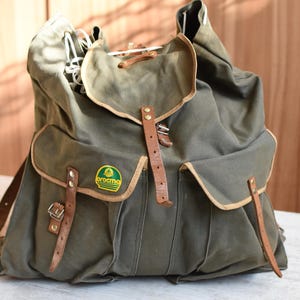 May include: A green canvas rucksack with brown leather straps and buckles. The rucksack has a large main compartment and two smaller pockets on the front. The rucksack has a logo that says "Brochma" on the front.