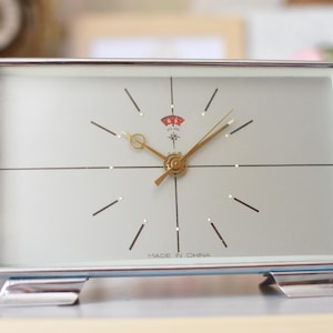 Op de afbeelding: Een rechthoekige analoge tafelklok met een zilverkleurig metalen frame en twee kleine voetjes. De wijzerplaat is lichtgrijs met dunne zwarte uurmarkeringen, goudkleurige wijzers, een centraal sterpictogram, een rode indicator en Made in China.