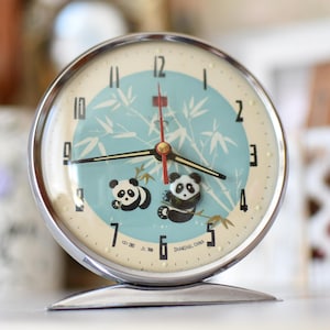 以下が含まれることがあります： 2 匹のパンダと竹のイラストが描かれた、ライトブルーの文字盤のヴィンテージクロム目覚まし時計。時計には黒い針、赤い秒針、シルバーのベースが付いています。文字盤には黒い数字があります。
