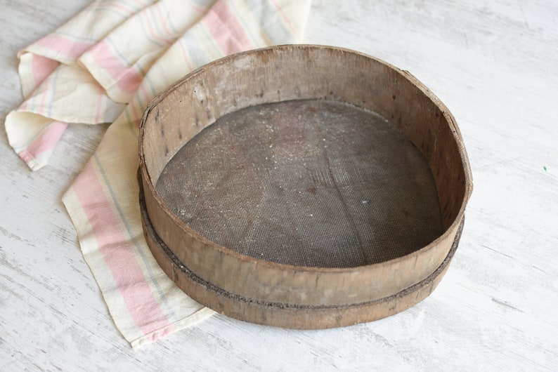 May include: A round wooden sifter with a wire mesh bottom. The sifter is made of natural wood and has a rustic, vintage look.