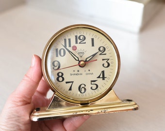 May include: A gold-colored desk clock with black Roman numeral markings and a diamond logo. The clock face reads "SHANGHAI CHINA".