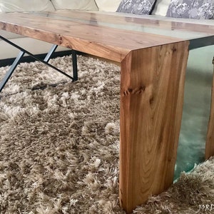 May include: A wooden coffee table with a clear epoxy resin inlay. The table has a natural wood finish and a black metal base.