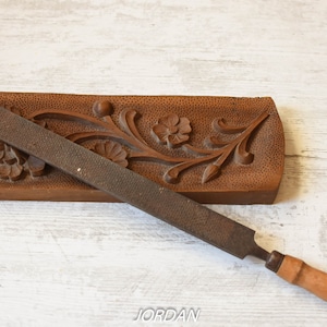 May include: A brown, wooden, hand-carved floral design with a metal rasp file. The file has a wooden handle. The floral design is on a rectangular wooden base.