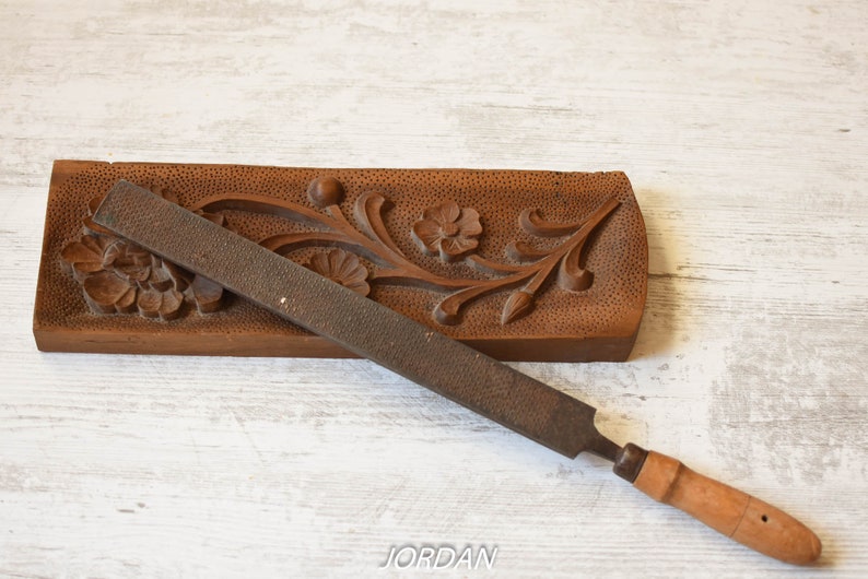 May include: A brown, wooden, hand-carved floral design with a metal rasp file. The file has a wooden handle. The floral design is on a rectangular wooden base.