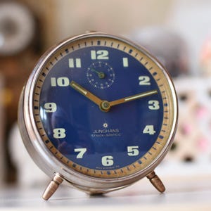May include: A round, silver-toned metal alarm clock with a deep blue face. The clock features gold-colored hands, light-colored Arabic numerals, and a small sub-dial. The text "JUNGHANS trivox-silentic" and "MADE IN GERMANY" are printed on the face.