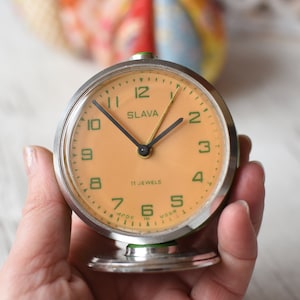 Puede incluir: Un reloj despertador vintage de cara naranja con números y manecillas verdes. La esfera del reloj dice "SLAVA" y "11 JEWELS".
