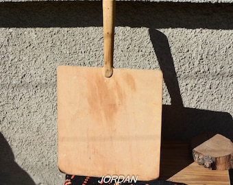 May include: A wooden pizza peel with a long handle. The peel is rectangular and has a light brown color. The handle is made of wood and is attached to the peel with a metal piece. The peel is sitting on a table with a wooden surface.