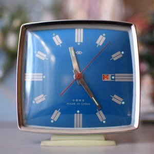 Könnte beinhalten: Ein quadratischer Vintage-Wecker mit blauem Zifferblatt und silbernen Zeigern. Die Uhr hat einen silbernen Rahmen und einen hellfarbigen Sockel. Das Zifferblatt zeigt weiße rechteckige Markierungen und den Text "MADE IN CHINA".